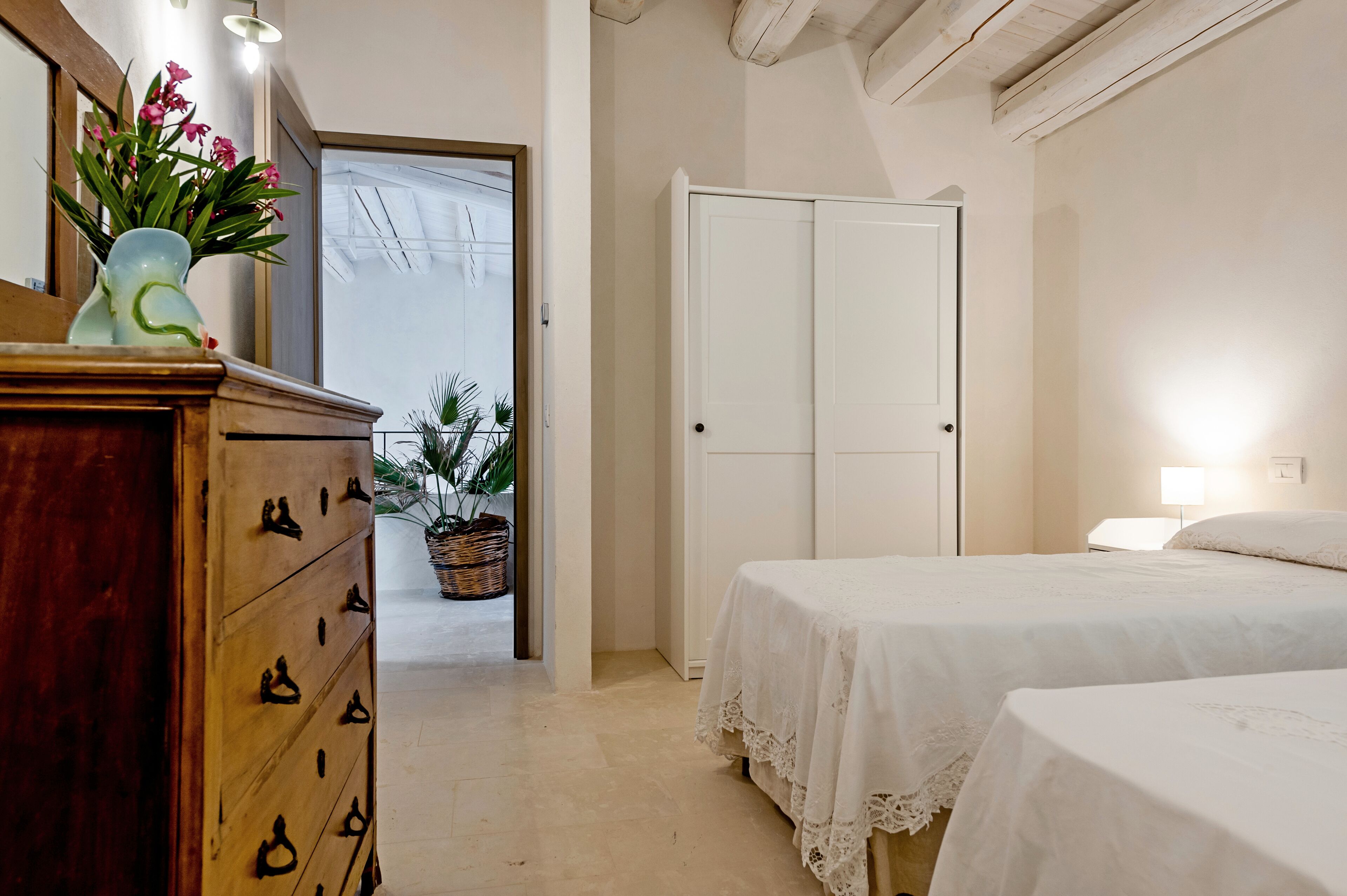 Een slaapkamer met twee bedden, een houten dressoir, een vaas met bloemen en een witte kledingkast.