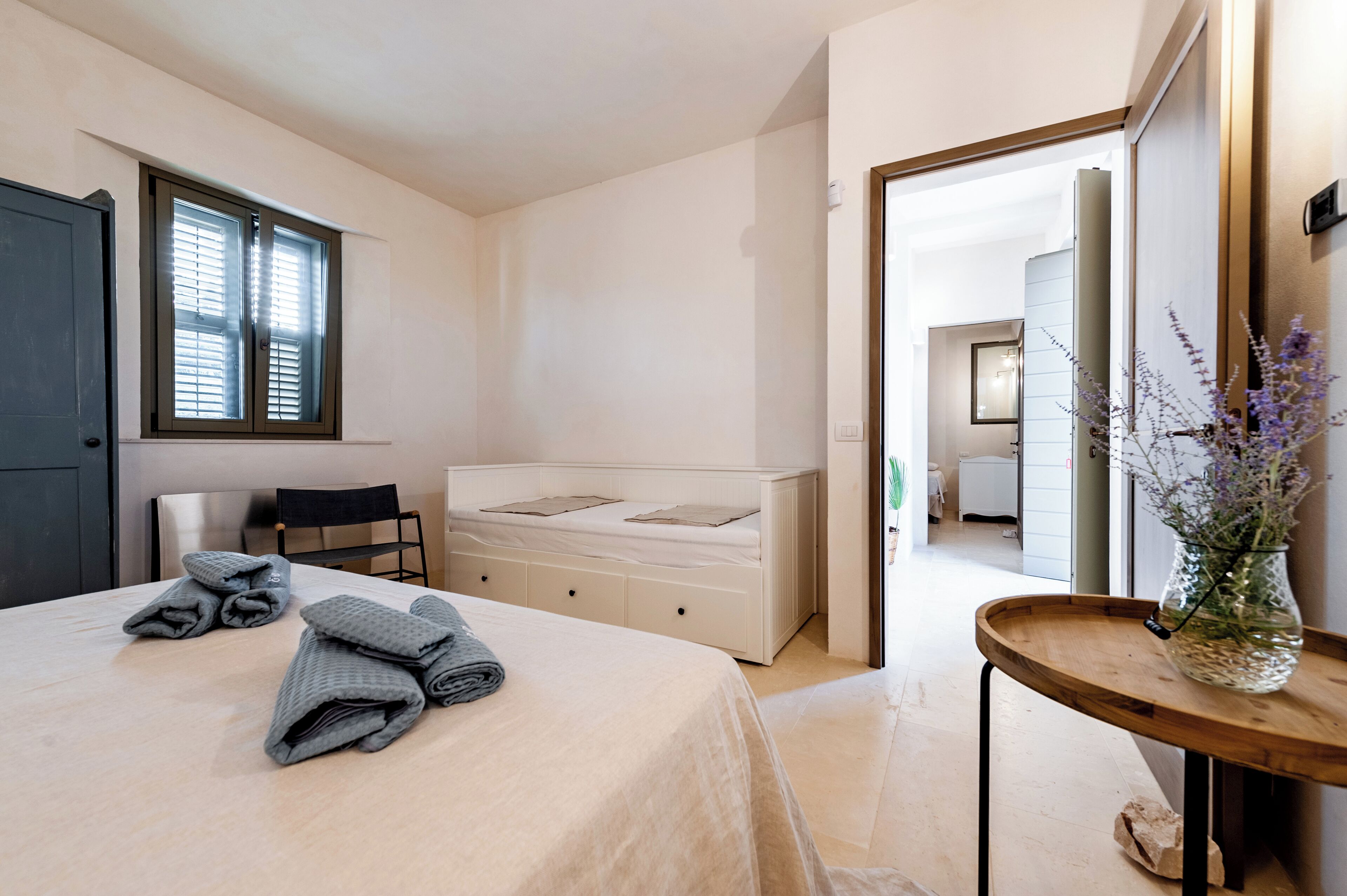 A bedroom with a bed, a chair, a bedside table with a vase, and a window with blinds.