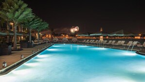 A nighttime swimming pool with lounge chairs and umbrellas, surrounded by buildings with warm lighting. - Stunning 4 Bd Close to Disney Magic Village Resort 7537 (Kissimmee)