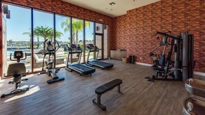 A gym with exercise equipment, a bench, and large windows overlooking a pool and palm trees. - Stunning 4 Bd Close to Disney Magic Village Resort 7537 (Kissimmee)
