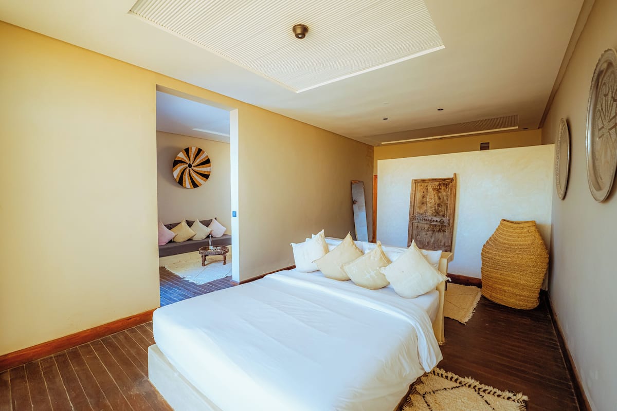 Een slaapkamer met een groot bed, een houten vloer, een plafondlamp en een deur naar een andere kamer.