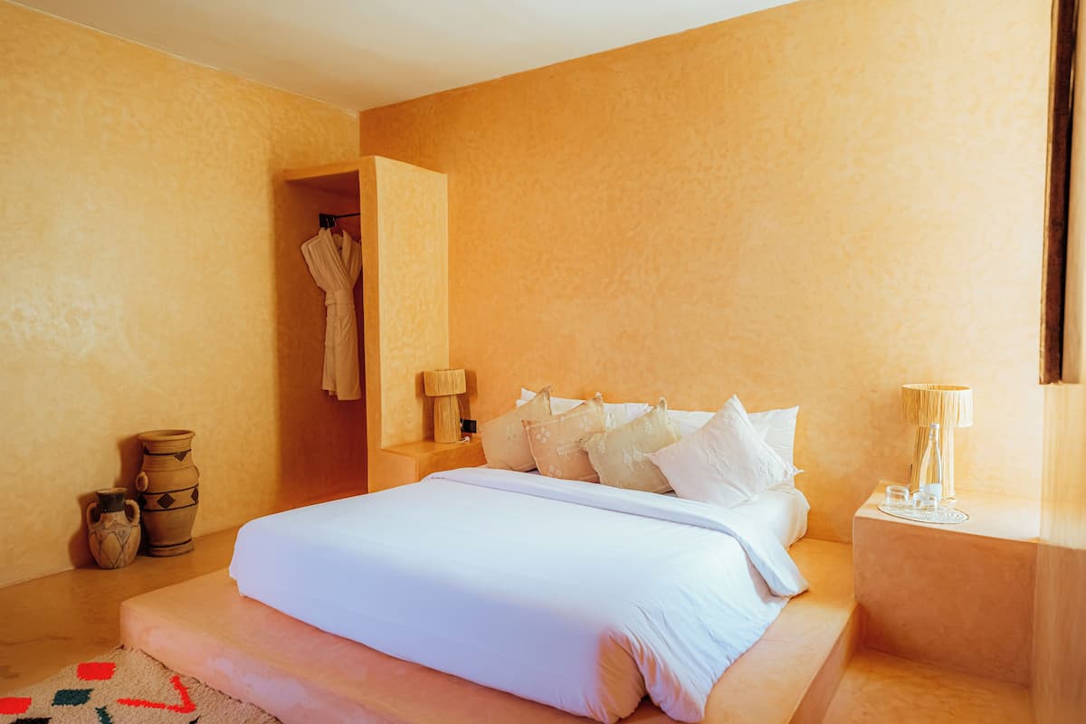 Een slaapkamer met een bed, kussens, een nachtkastje, een lamp en een vaas.