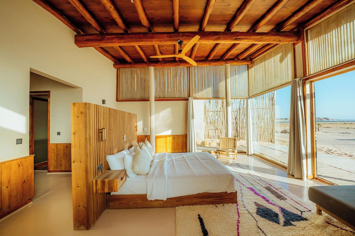 Een slaapkamer met een houten plafond, een bed met witte lakens, een groot raam met gordijnen en uitzicht op het strand.