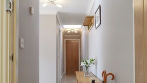 Un pasillo con un banco de madera, un florero con flores blancas y un cubo de metal con paraguas.
