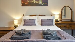 Una cama bien hecha con dos almohadas, una mesita de noche con una lámpara, un aparador de madera con un espejo y un cuadro de un paisaje urbano en la pared.