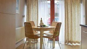 Un comedor con una mesa redonda, cuatro sillas de mimbre, una botella de vino y vistas a través de una ventana con cortinas florales.