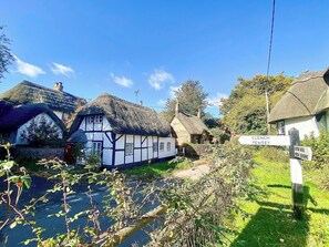 Een traditioneel vakantiehuis met rieten dak, een wit en zwart vakwerkstructuur, een bord met 'Clench Pewsey' en een helderblauwe lucht.