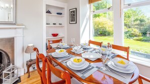 Ein Essbereich mit einem Tisch für vier Personen, einem Kamin und Blick auf einen Garten durch große Fenster.