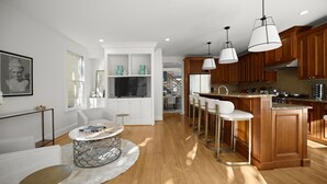 Una cocina moderna con armarios de madera, una isla central con taburetes, un comedor con una mesa redonda y un televisor en la sala.