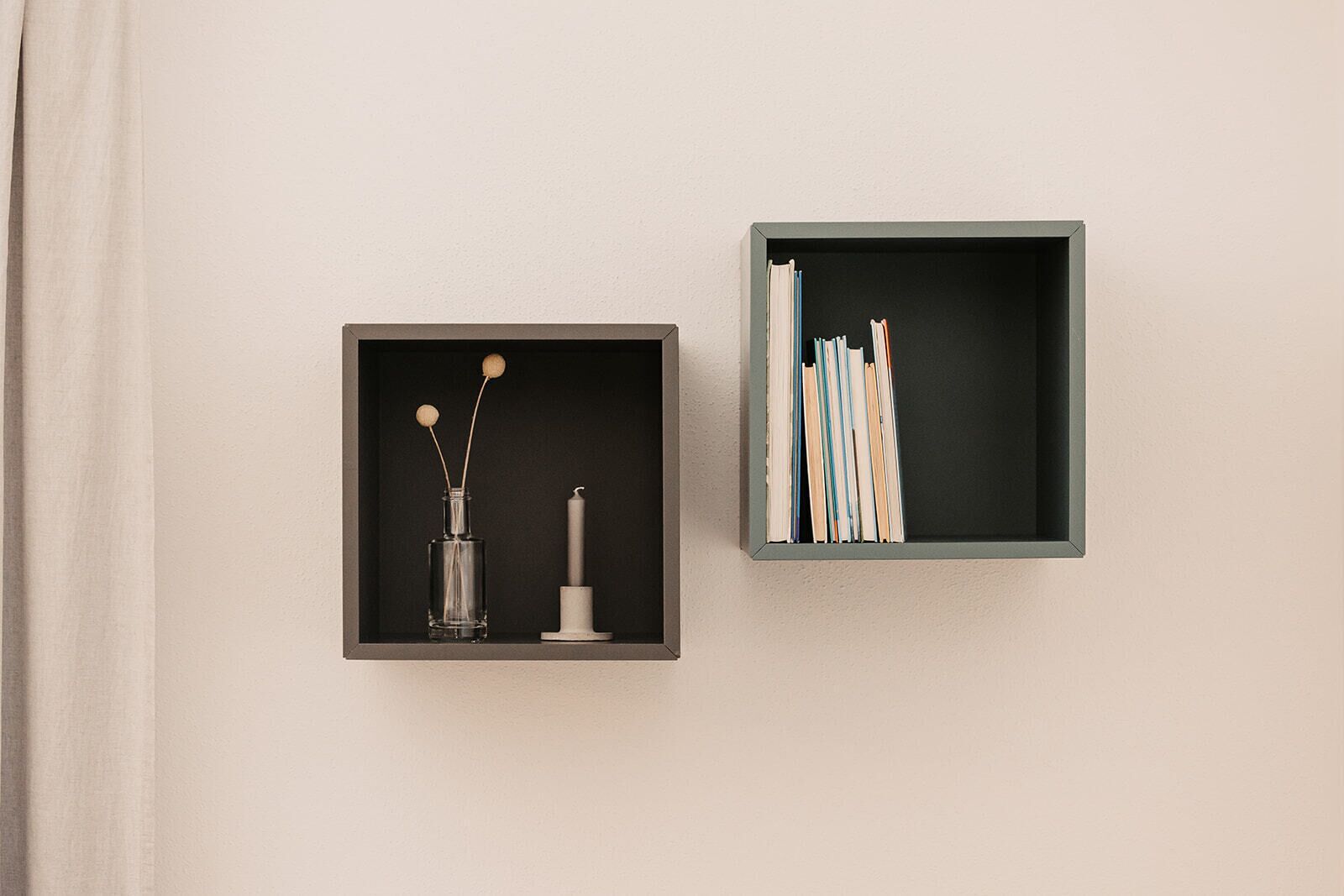 Two square shelves against a wall, one with a clear glass bottle and a candle, the other with books.