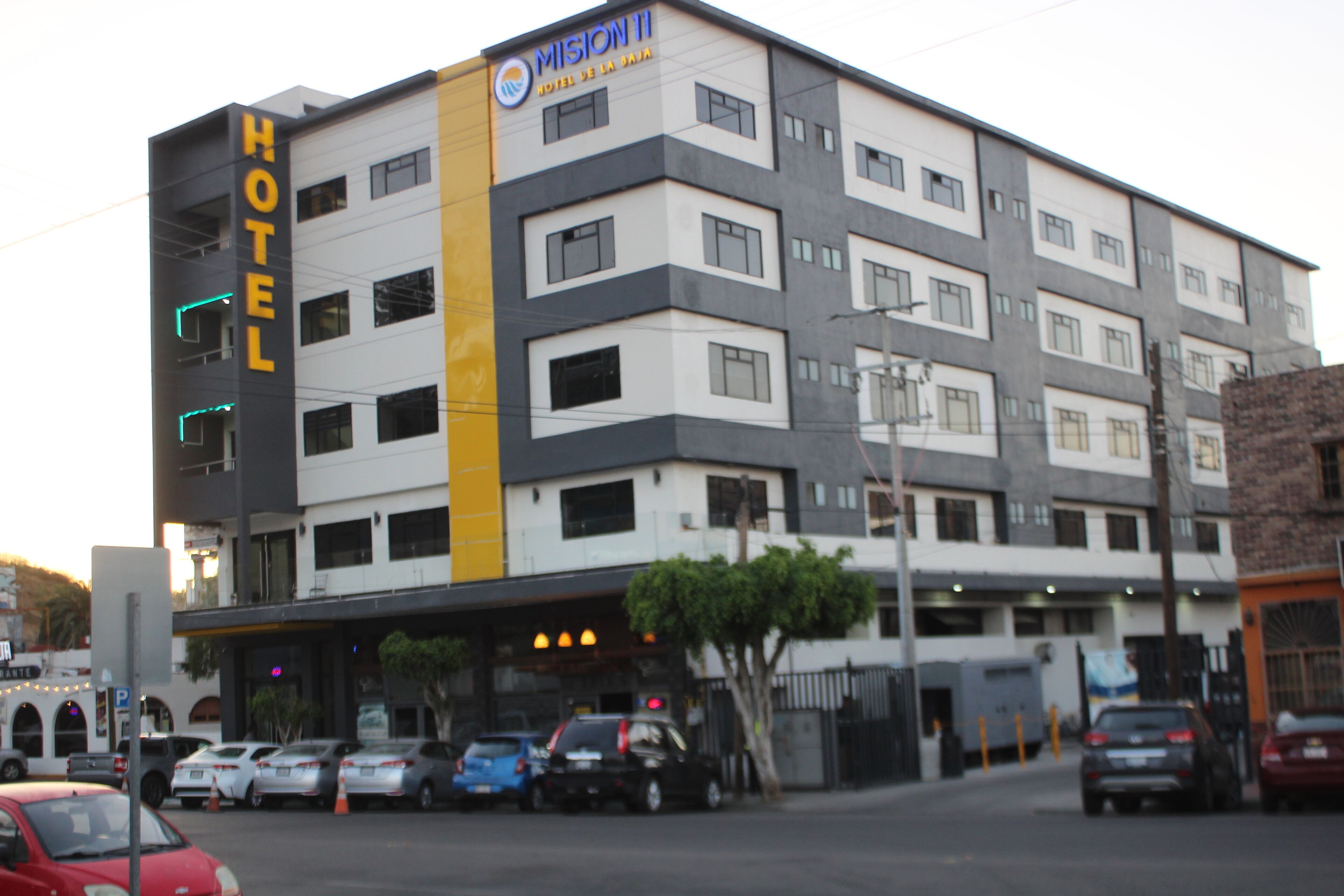 A multi-story hotel with a prominent 'HOTEL' sign on the facade, a parking lot with several cars, and a tree in front of the entrance.
