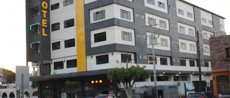 Un hotel de varios pisos con un cartel grande que dice 'HOTEL' en la fachada, un estacionamiento con varios coches y un árbol frente a la entrada.