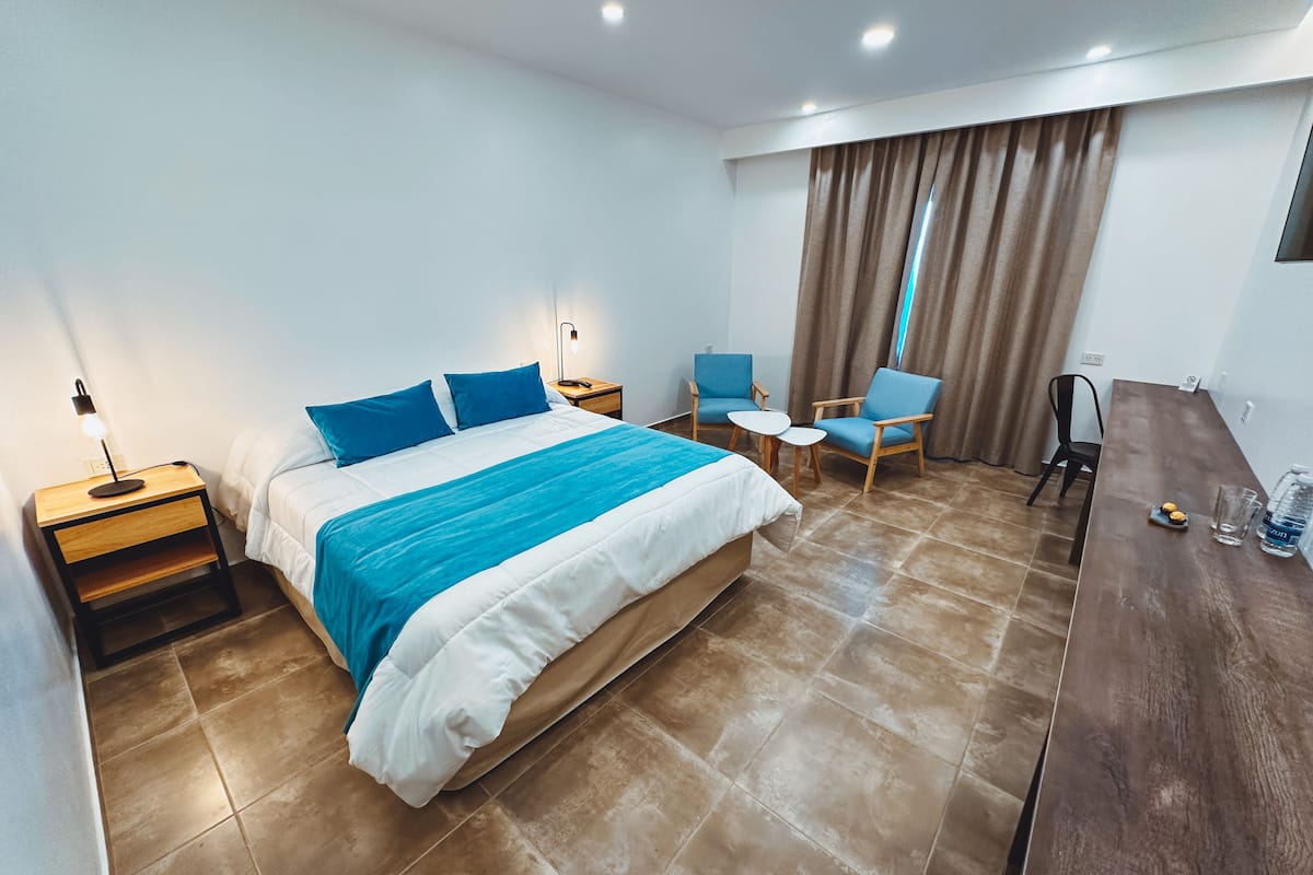 A hotel room with a large bed, two bedside tables, a desk with chairs, a small table, and a window with curtains.