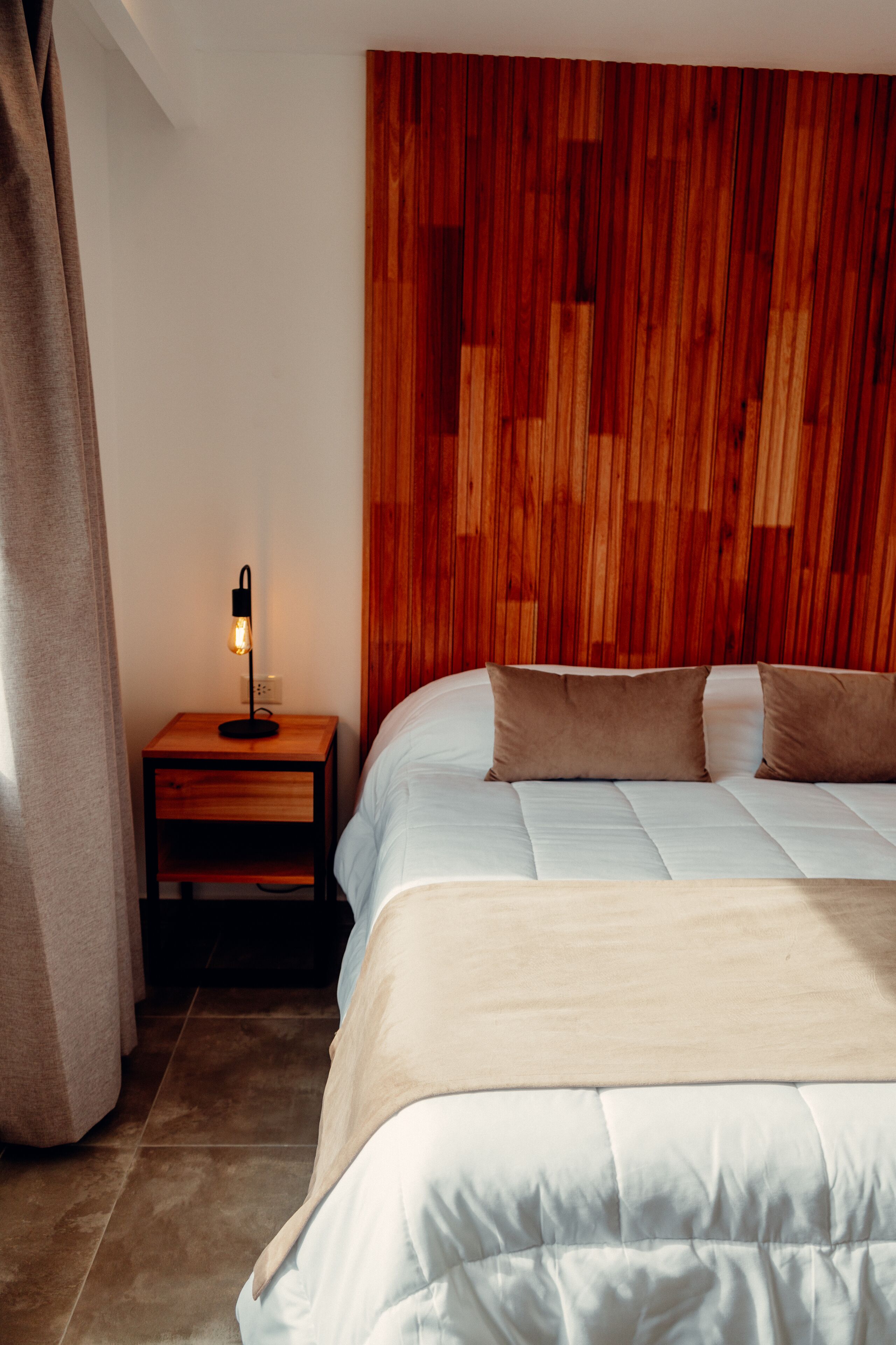 Ein Schlafzimmer mit Holz-Kopfteil, einem Nachttisch mit Lampe, einem Bett mit weißer Bettwäsche und einem Vorhang auf der linken Seite.