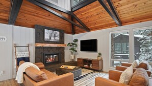 A cozy living room with a fireplace, wooden ceiling beams, and a large window overlooking a snowy landscape. - 12 Quartz Mtn - Updated and Classy! Pickleball, SHARC, Fireplace, Backs to Sunriver Forest (Sunriver)