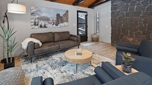 A modern living room with a brown leather sofa, a round wooden coffee table, and a stone wall. - 12 Quartz Mtn - Updated and Classy! Pickleball, SHARC, Fireplace, Backs to Sunriver Forest (Sunriver)