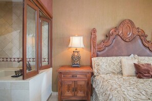 A bedroom with a wooden nightstand, a lamp, a bed with patterned bedding, and a mirror above the nightstand.
