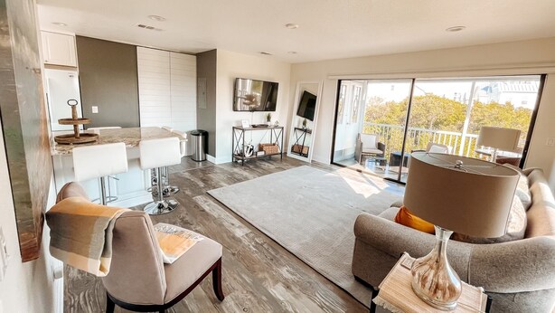 A modern living room with a large window, a flat-screen TV, and a dining area with a kitchen counter. - Welcome to Paradise Cove! - Steps From The Beach (Indian Shores)