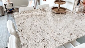 A modern dining area with a large granite table, white upholstered chairs, and a wooden serving tray. - Welcome to Paradise Cove! - Steps From The Beach (Indian Shores)