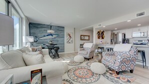 En moderne stue med sofa, lænestole, et sofabord og et tæppe. Der er et spisebord med stole, et køkken med hårde hvidevarer, og et stort vindue.
