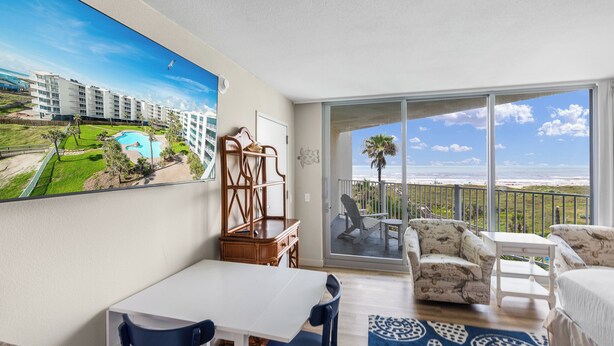 A balcony with a view of a pool and buildings, a dining area with a table and chairs, and a patterned rug.