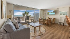 A spacious living room with a large sliding glass door leading to a balcony with a view of the ocean. There is a dining area with wicker furniture and a flat-screen TV mounted on the wall.