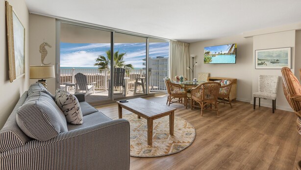 A spacious living room with a large sliding glass door leading to a balcony with a view of the ocean. There is a dining area with wicker furniture and a flat-screen TV mounted on the wall.