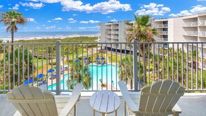 A balcony with three Adirondack chairs and a small table, overlooking a swimming pool and beach area.