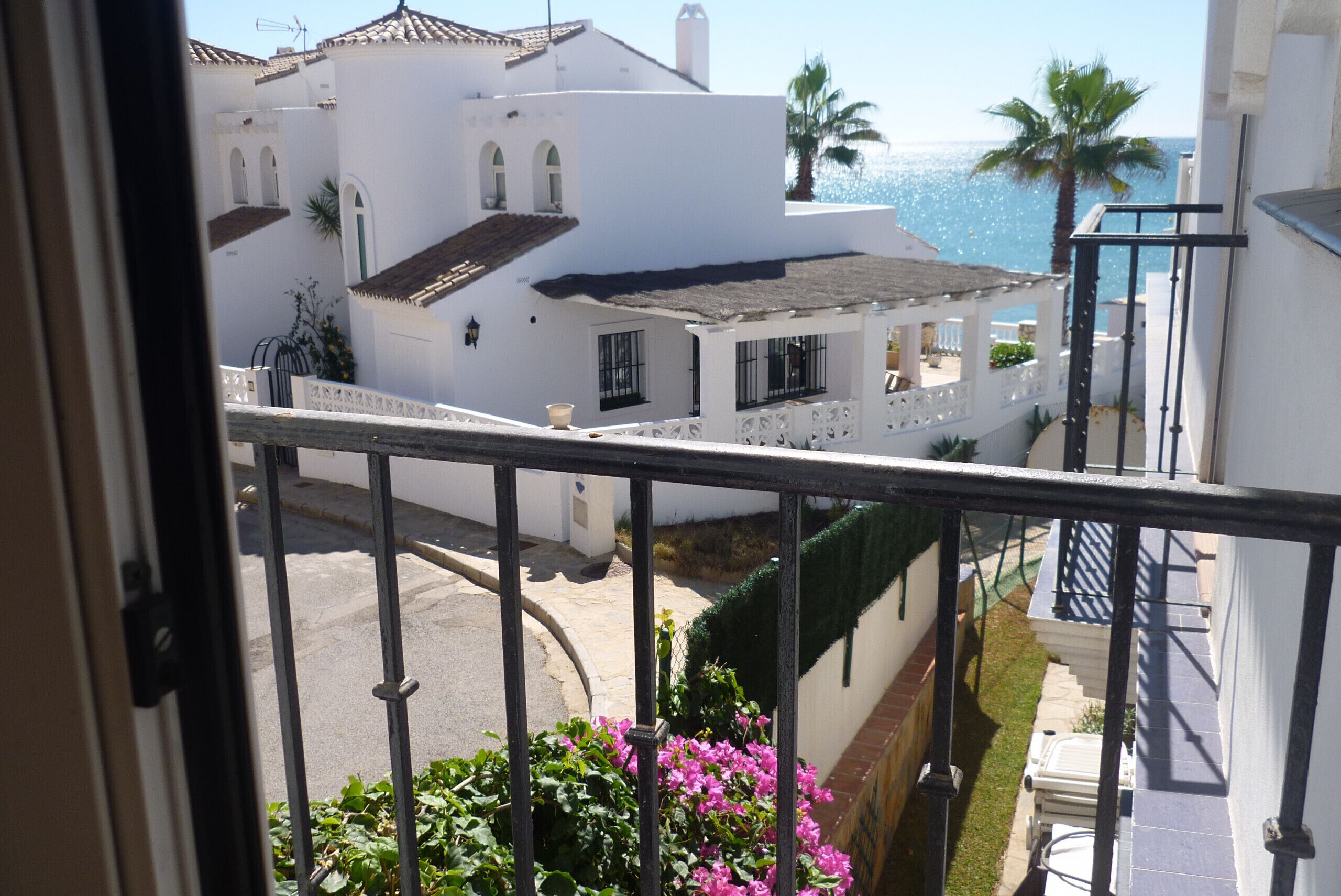 Uma varanda com grades de metal, vista para uma rua com edifícios brancos, uma palmeira e o mar.