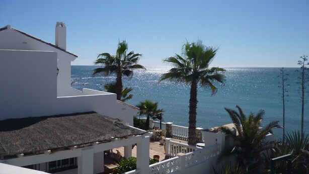 A coastal building with a white facade, a thatched roof, and a balcony overlooking the sea.
