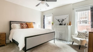 Un dormitorio con cama, ventilador de techo, chimenea y ventana con vista a edificios.