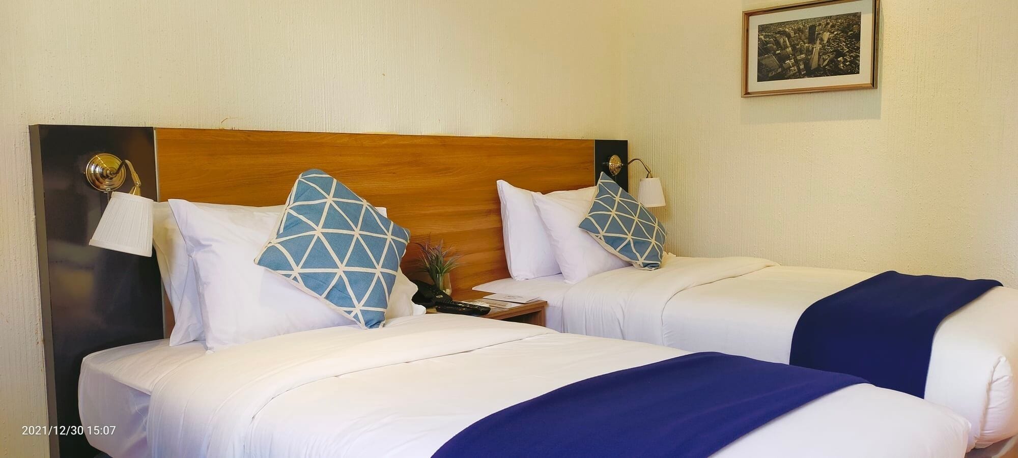 Habitación de hotel con dos camas, cada una con ropa de cama blanca y almohadas azules geométricas. Tiene un cabecero de madera, una mesita de noche con lámpara y un cuadro en la pared.