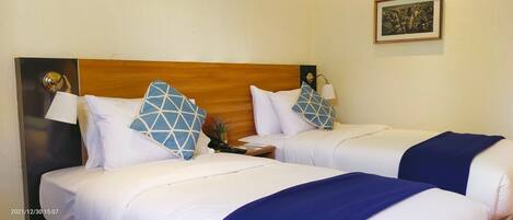 Habitación de hotel con dos camas, cada una con ropa de cama blanca y almohadas azules geométricas. Tiene un cabecero de madera, una mesita de noche con lámpara y un cuadro en la pared.