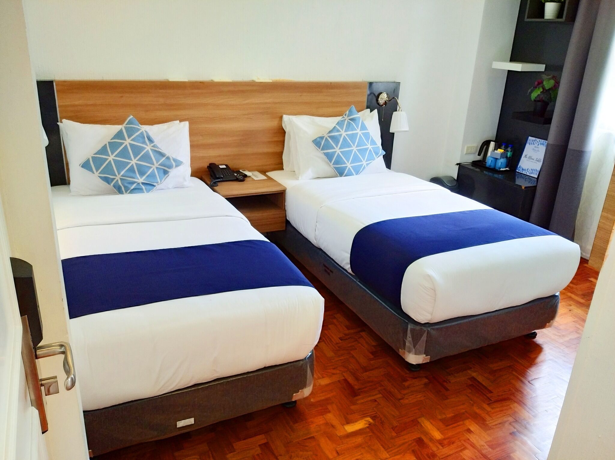 A hotel room with two beds, each with a wooden headboard, white bedding, and blue accents. There is a bedside table with a lamp and a phone.