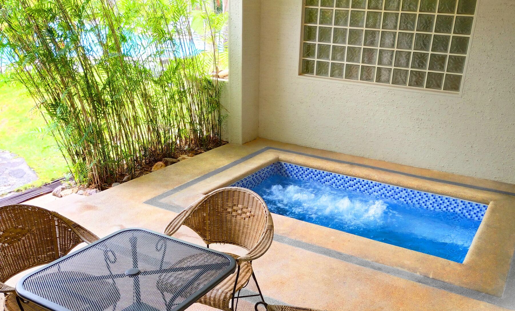 A small outdoor hot tub area with a tiled floor, two wicker chairs, and a glass door leading to a garden with bamboo plants.