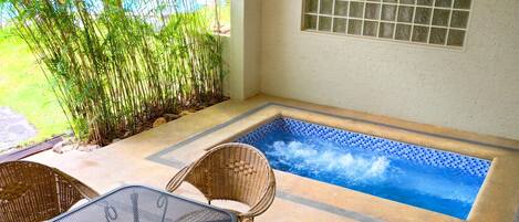 A small outdoor hot tub area with a tiled floor, two wicker chairs, and a glass door leading to a garden with bamboo plants.