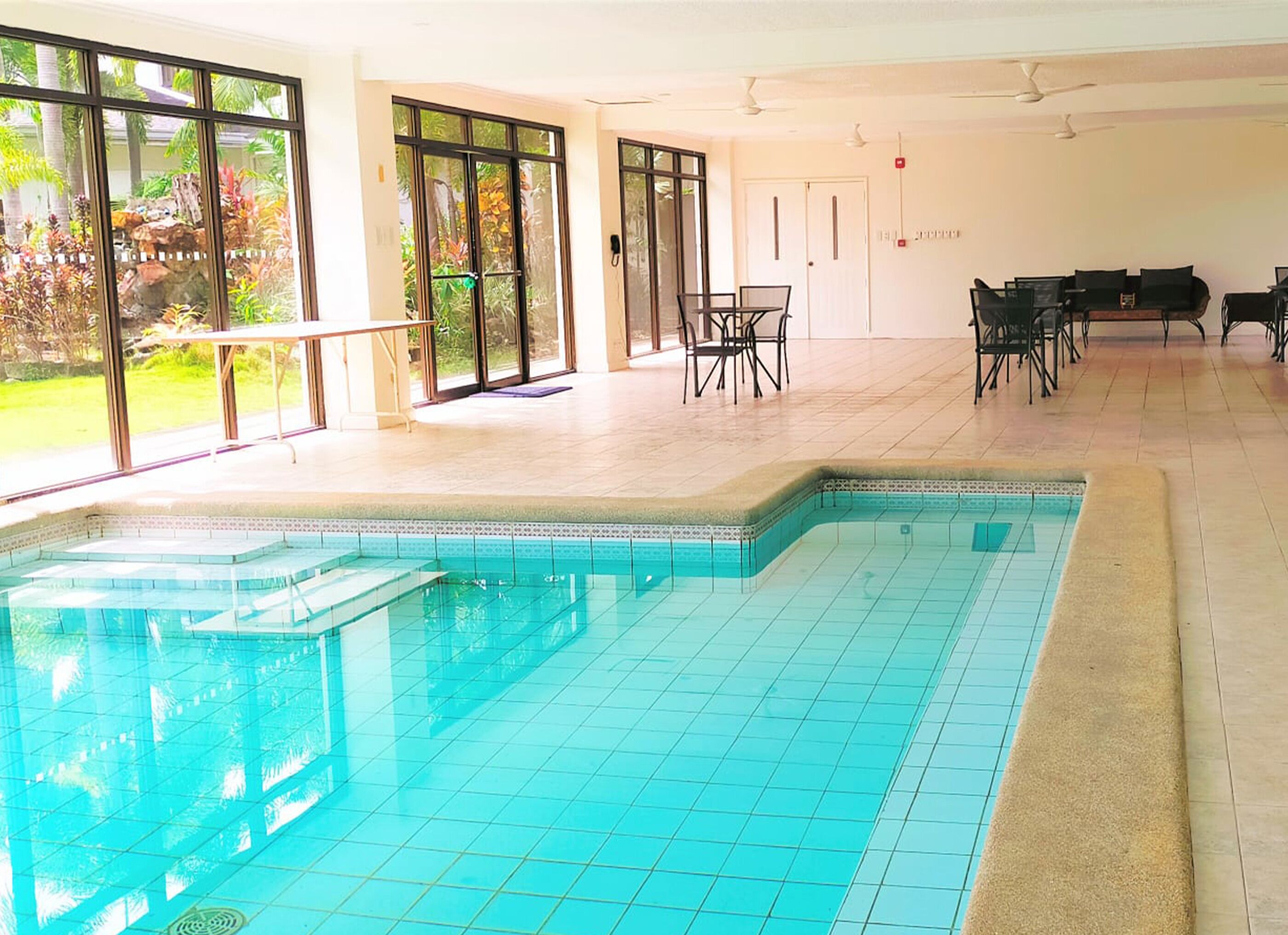 A spacious indoor swimming pool with clear blue water, surrounded by large windows offering views of a lush garden.