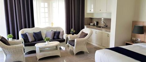 Habitación de hotel con cama, sofá, dos sillones, una mesita, zona de cocina y ventana con cortinas.