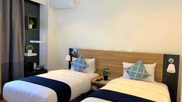 Habitación de hotel con dos camas, una mesita de noche con lámpara, un pequeño estante con una planta y un aire acondicionado instalado en la pared.