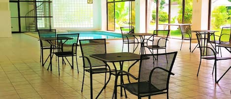 A spacious indoor area with tiled flooring, multiple tables and chairs, a pool, and large windows offering views of greenery.