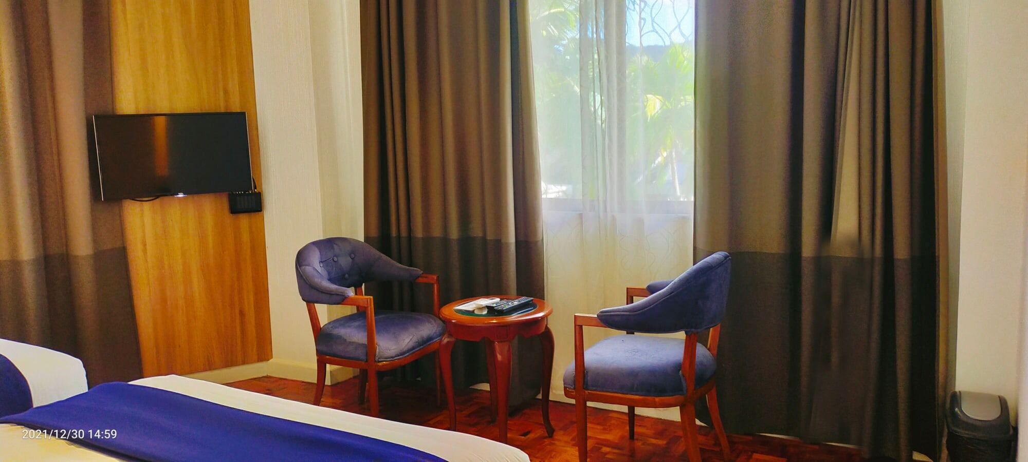 A hotel room with two blue upholstered chairs, a small round table with a remote control, and a flat-screen TV mounted on the wall.