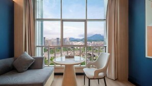 Quarto com sofá, mesa redonda, cadeira, janela grande com vista para edifícios e uma montanha, e parede azul.