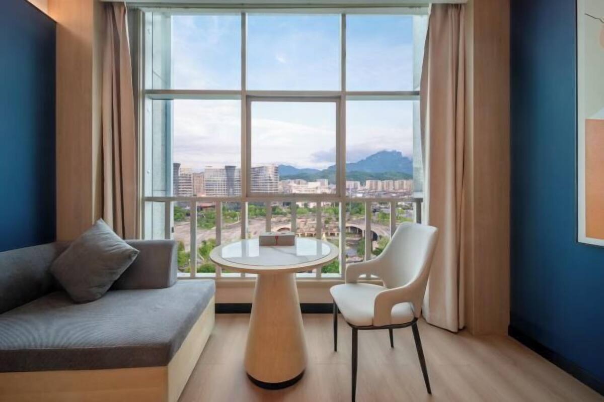 Ein Zimmer mit einer Couch, einem runden Tisch und einem Stuhl, ein großes Fenster mit Blick auf Gebäude und einen Berg, sowie eine blaue Wand.