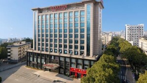 Um hotel moderno de vários andares com um letreiro chamativo da marca RAMADA, fachada de vidro e estacionamento em frente.