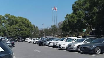 Um estacionamento com vários carros estacionados em filas, árvores ao longo da rua e bandeiras em mastros ao fundo.