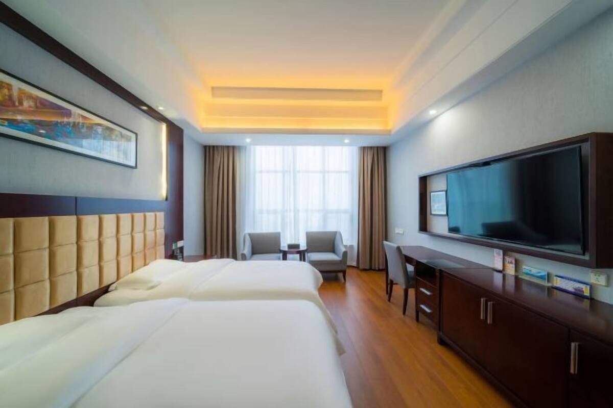 Ein Hotelzimmer mit zwei Betten, einem Fernseher, einem Schreibtisch und einem Fenster mit Vorhängen.
