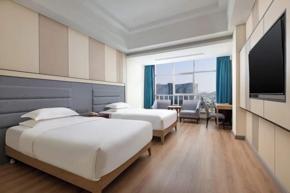 Une chambre d’hôtel avec deux lits, un bureau, une chaise, une télévision et une grande fenêtre avec des rideaux.