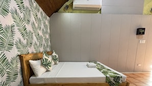 Una cama de madera con colchón blanco y almohadas estampadas con hojas, frente a una pared con un diseño similar de hojas.