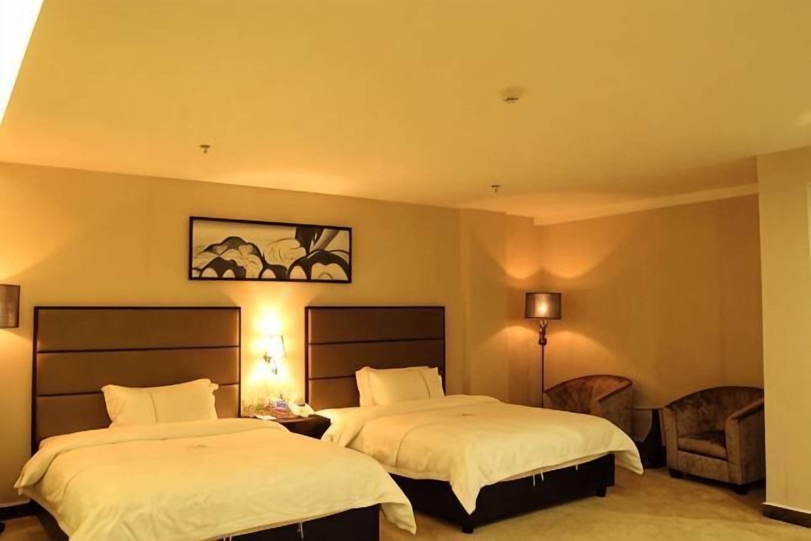 Habitación de hotel con dos camas, una mesita de noche, una lámpara, una silla y un cuadro en la pared.