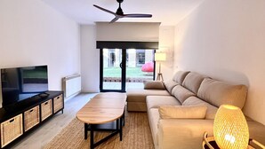 A modern living room with a flat-screen TV, a wooden coffee table, a beige sofa, a standing lamp, and a ceiling fan.